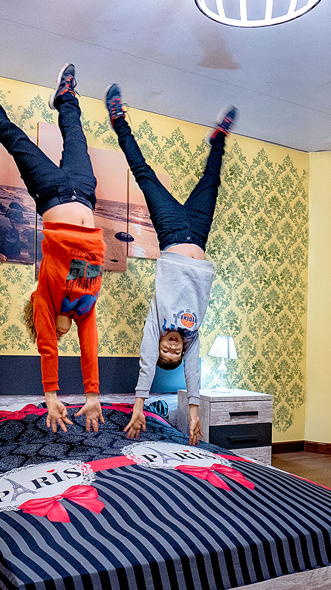 Deux enfants font le poirier à l'envers sur un lit rayé aux couleurs de Paris dans une chambre inversée à Upside Down House Barcelona