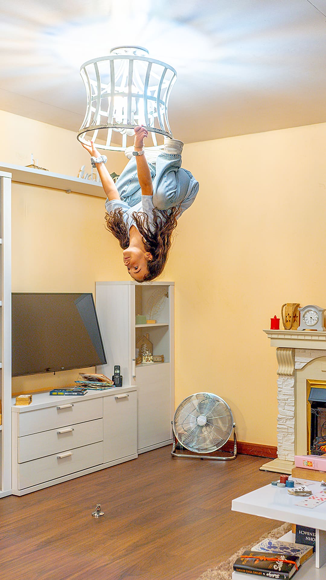 Une femme suspendue à l'envers à un luminaire de plafond au-dessus d'un salon inversé avec cheminée et télévision à Upside Down House Barcelona
